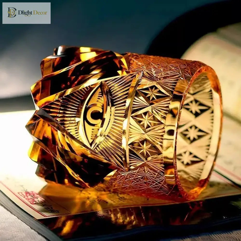 Amber-gold crystal tumbler showing engraved eye design and geometric facets along the side