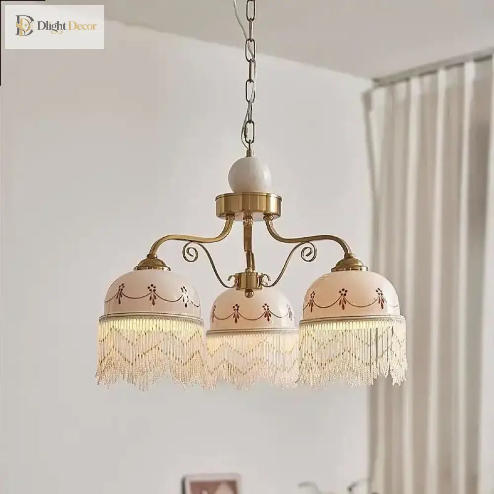 Gold three-head pendant light with white ceramic shades, brown floral trim, fringe tassels on chain