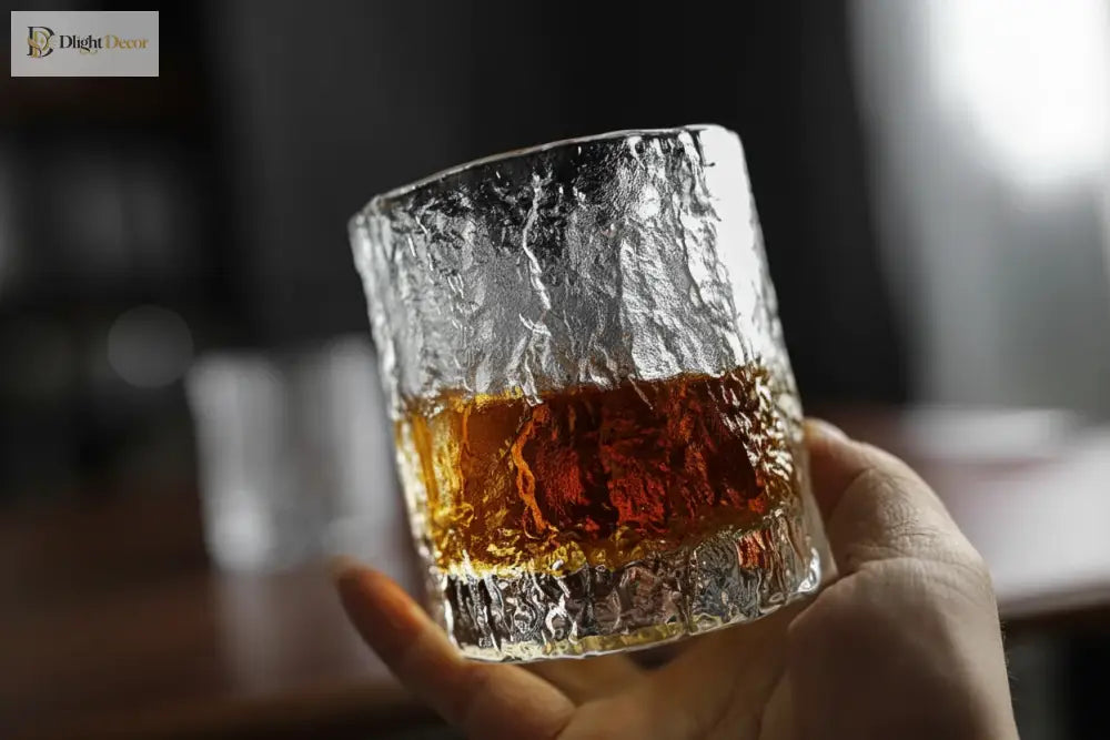 Hammered crystal rocks glass held by hand, with amber drink inside.