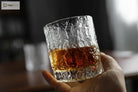 Hammered crystal rocks glass held by hand, with amber drink inside.