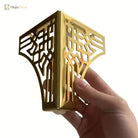 Gold-tone metal furniture leg corner bracket with geometric cutouts, held in hand