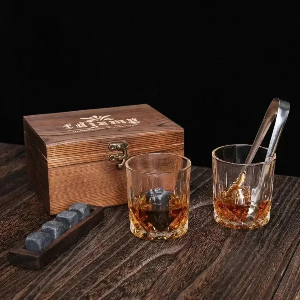 Wooden whiskey box with two tumblers, chilling stones, and metal tongs on wood surface
