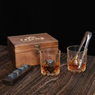 Wooden whiskey box with two tumblers, chilling stones, and metal tongs on wood surface